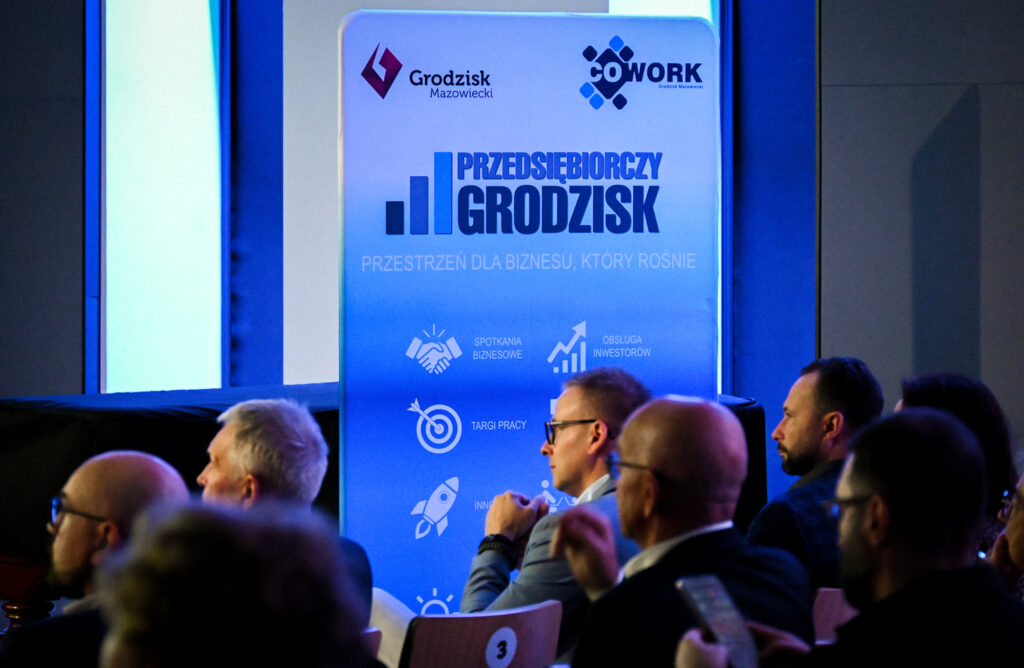 Przedsiębiorcy i prelegenci siedzą na krzesłach przy banerze Przedsiębiorczego Grodziska.