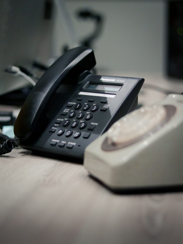 Dwa telefony na biurku z czego jeden jest biały a drugi czarny. Są to telefony stacjonarne.